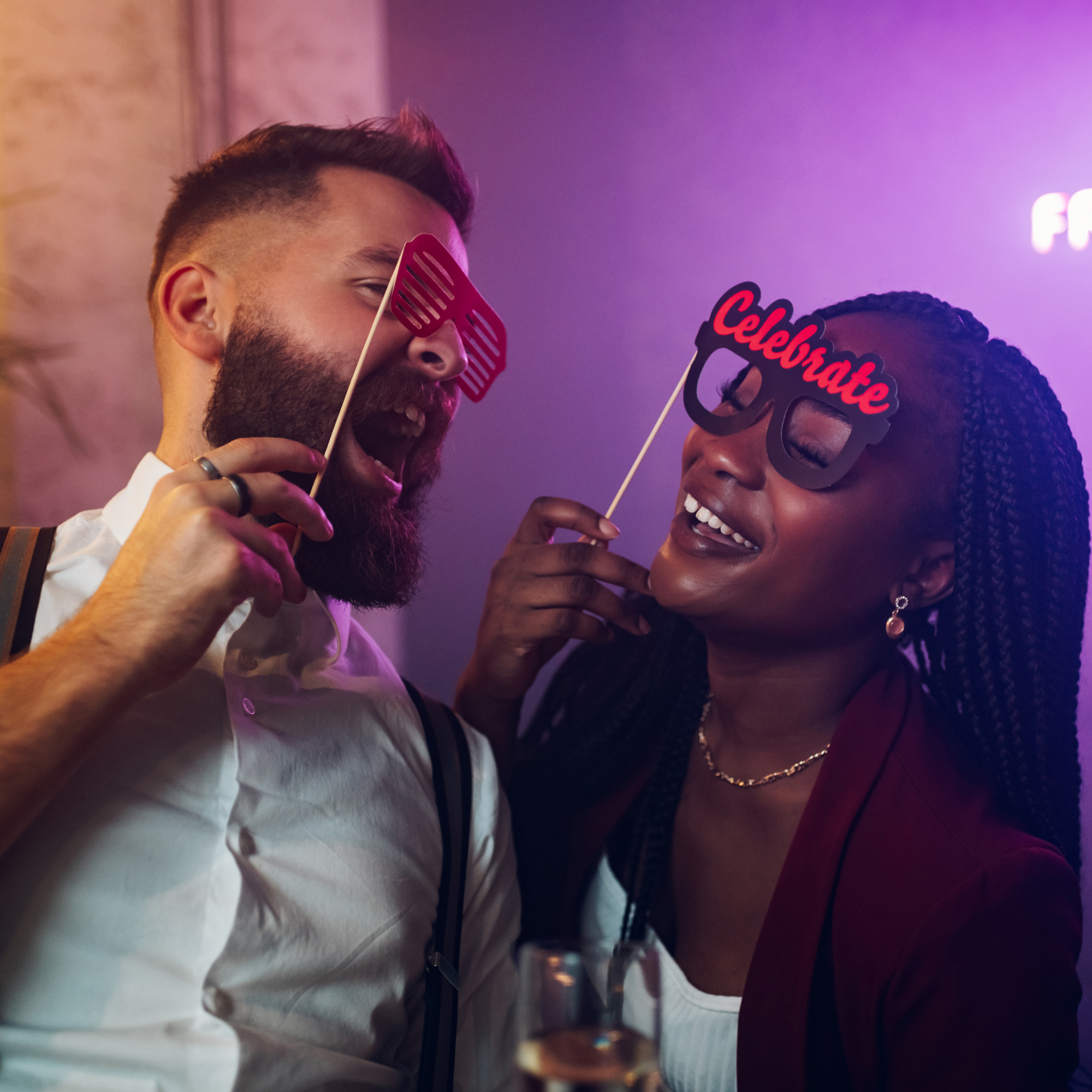 A candid photo of a man and woman laughing and posing together, holding up novelty paper glasses as props in a dimly lit, festive setting with neon purple and pink lights, conveying high energy and fun at an event.