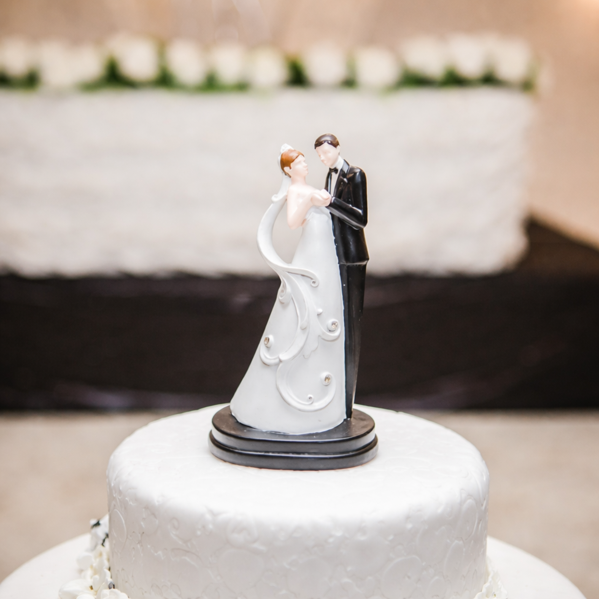 Close-up of a bride and groom wedding cake topper on a white textured cake with blurred floral background.